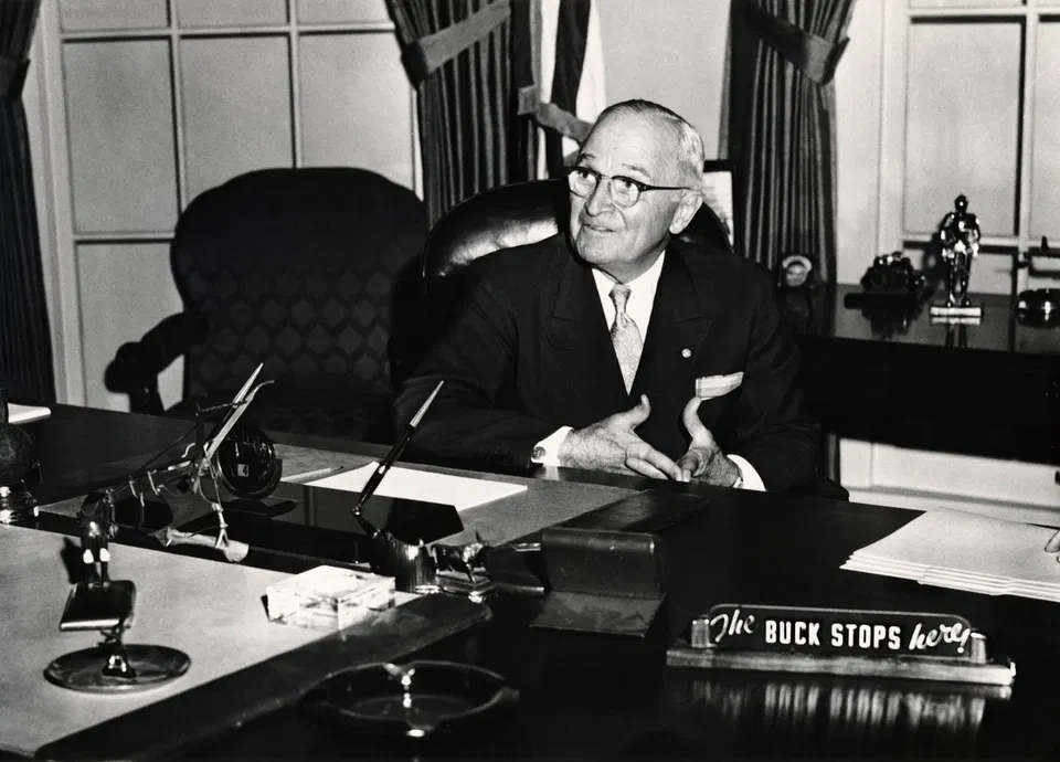 US President Harry S. Truman at his desk beside a sign reading “The Buck Stops Here”.